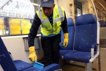 Schoonmaker met gele handschoenen en veiligheidsvest die de stoelen in een trein schoonmaakt met een handborstel.