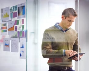 Man die op kantoor tegen glazen wand leunt en op tablet kijkt, links van hem hangen grafieken en post its op het glas
