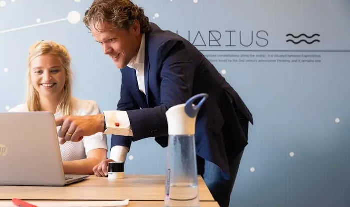 Een man staat naast een zittende vrouw en wijst naar het scherm van een laptop op een bureau, terwijl ze samen naar de inhoud kijken in een moderne kantooromgeving.