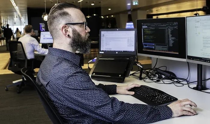 Man with glasses working behind three screens