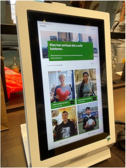 Touchscreenzuil met een interface waarop verschillende persoonlijke verhalen met foto’s van mensen worden getoond, geplaatst in een binnenruimte met tafels en bezoekers op de achtergrond.