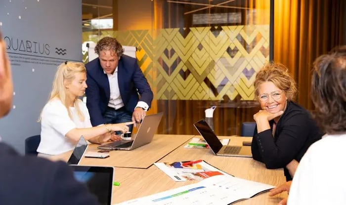 Groep collega’s zit rond een tafel in een vergaderruimte, waarbij twee personen samenwerken achter een laptop en een vrouw glimlachend naar de camera kijkt, met laptops en documenten op tafel.