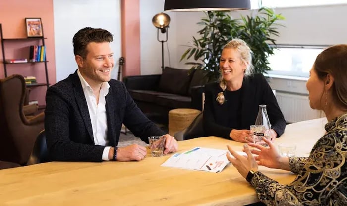 Drie mensen zitten rond een tafel in een informele vergaderruimte en lachen tijdens een gesprek, met documenten en glazen water op tafel.