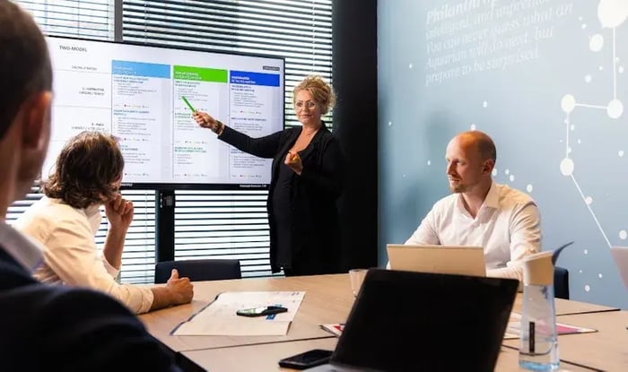Een vrouw geeft een presentatie in een vergaderruimte en wijst naar een scherm met schema’s, terwijl collega’s aan tafel luisteren en werken met laptops en notities.