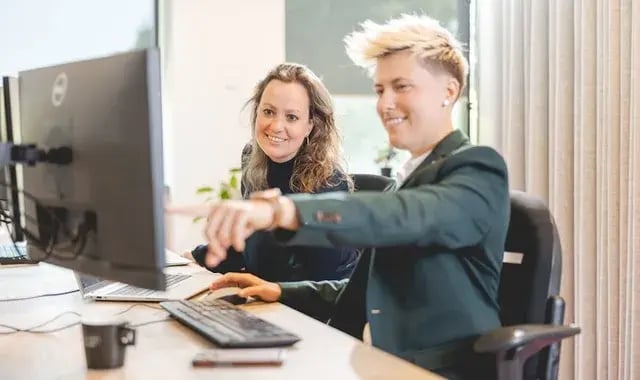 Twee collega's zitten naast elkaar en één wijst naar het computerscherm.
