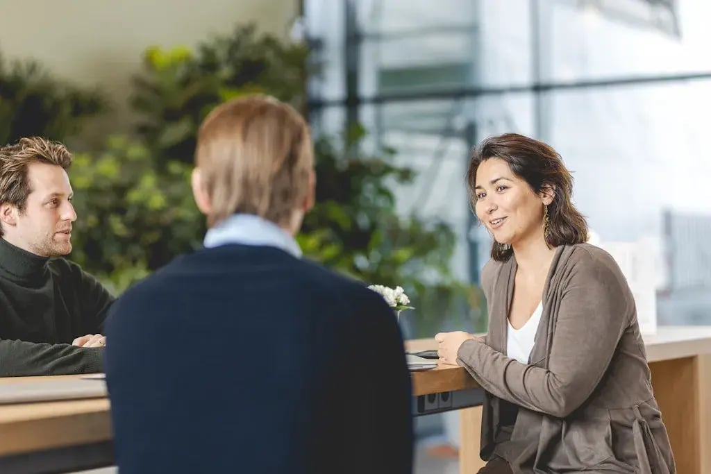 Drie mensen in een vergaderruimte, waarbij een vrouw met een glimlach aan het praten is met de twee mannen aan tafel.
