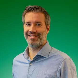 Portret van een glimlachende man met kort grijs haar en lichte baard, gekleed in een lichtblauw overhemd, tegen een groene achtergrond.
