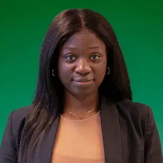 Portret van een vrouw met lang donker haar, gekleed in een zwart jasje en lichtgekleurde top, die recht in de camera kijkt tegen een groene achtergrond.