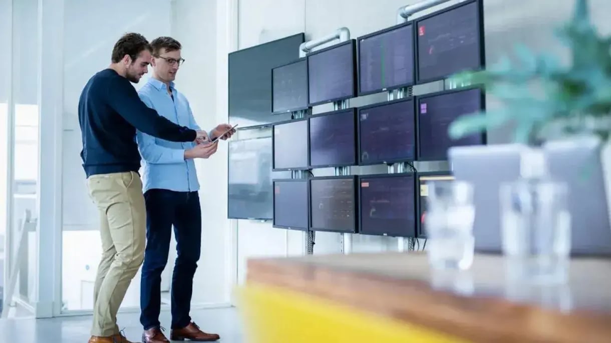 Two men talking in front of several screens 
