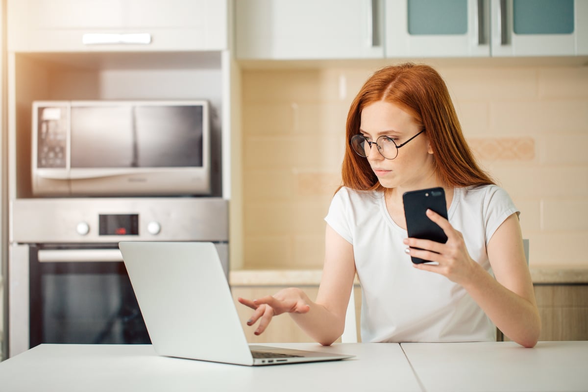 Vrouw die in de keuken werkt op een laptop en haar smartphone vasthoudt.
