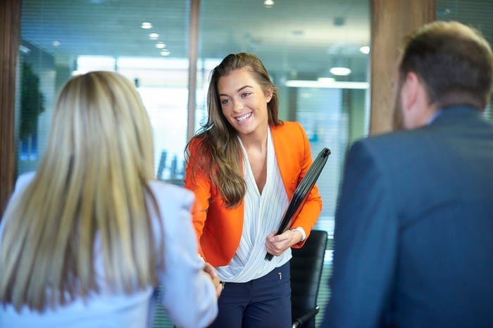 Een vrouwelijke collega in een oranje blazer schudt de hand met een andere vrouw.