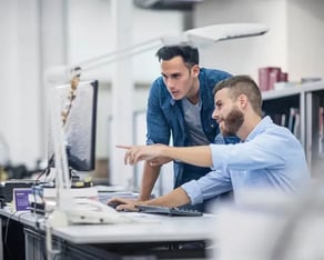 Man die naar zijn beeldscherm wijst en collega die over zijn schouder meekijkt