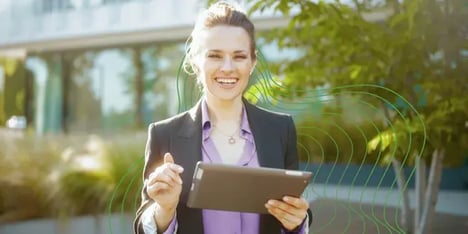 A white lady with dark hair in a ponytail with a purple shirt, smiling in front of the camera with a tablet in her hand.