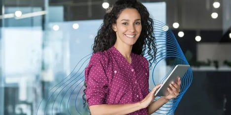 A white lady with brown curly long hair, smiling in front of the camera, in a magenta blouse with white dots and a tablet in her hand