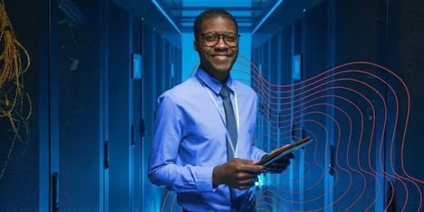 A darkskin man with glasses, smiling in front of the camera, in blue shirt with tie and a tablet in his hands