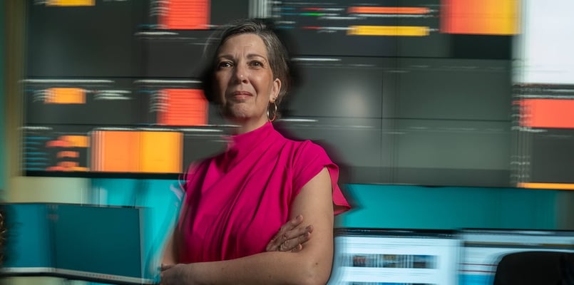 Vrouwelijke collega staat met armen over elkaar voor computerschermen.