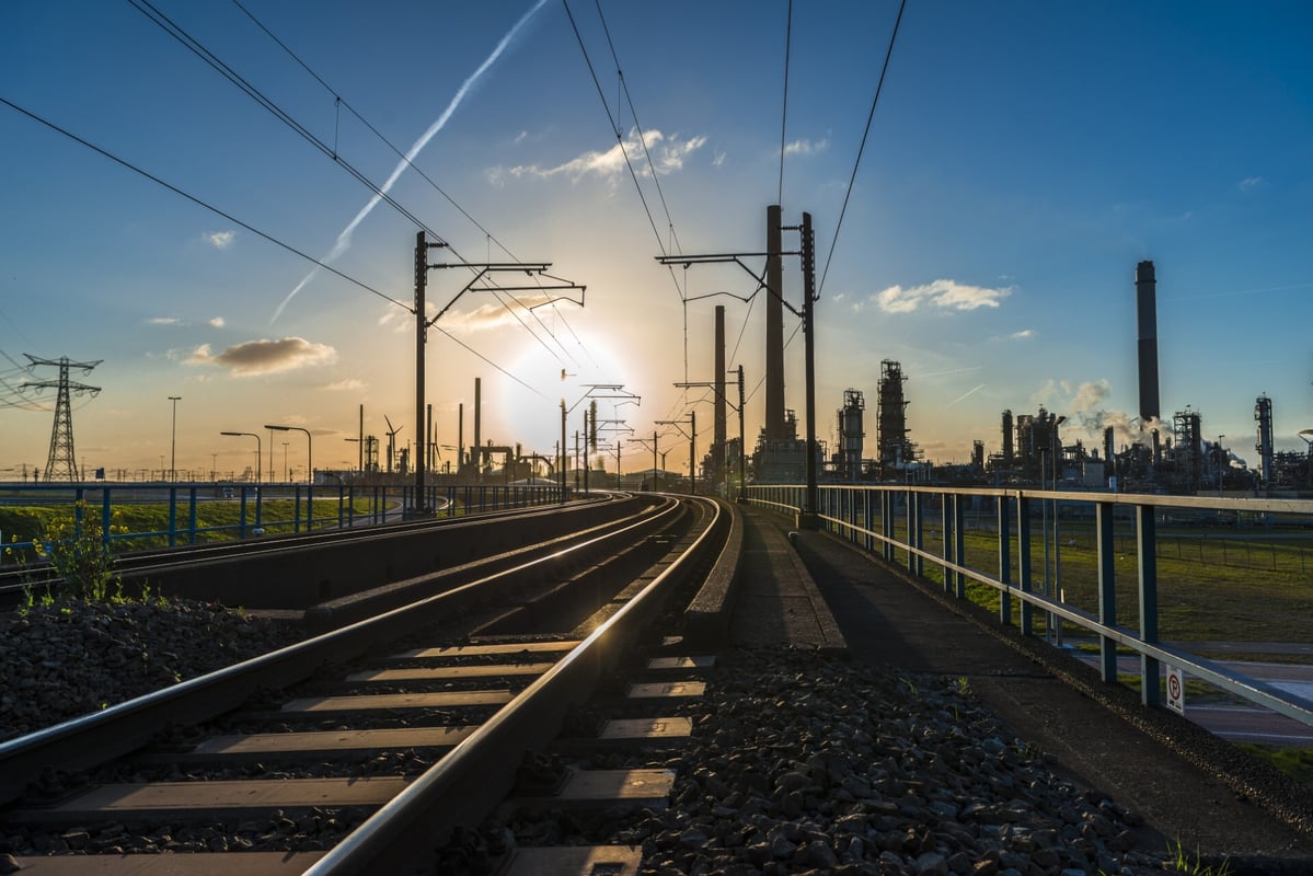 A train track runs through an industrial area where the sun can be seen rising in the distance with some factories next to it