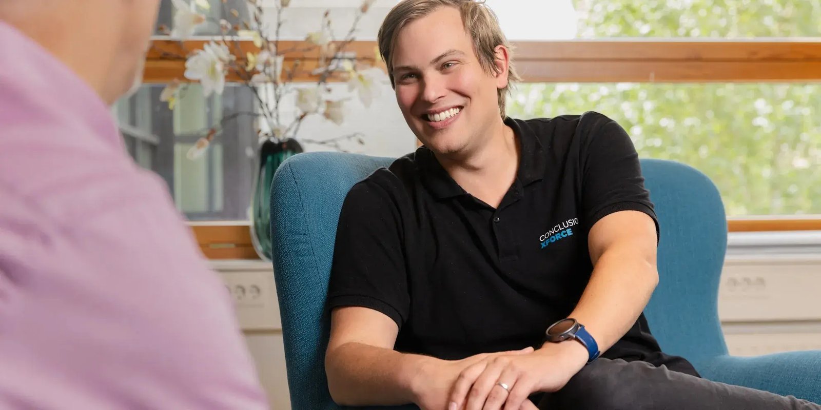 Mannelijke Xforce collega zit op een stoel en lacht vriendelijk naar de collega tegenover hem.