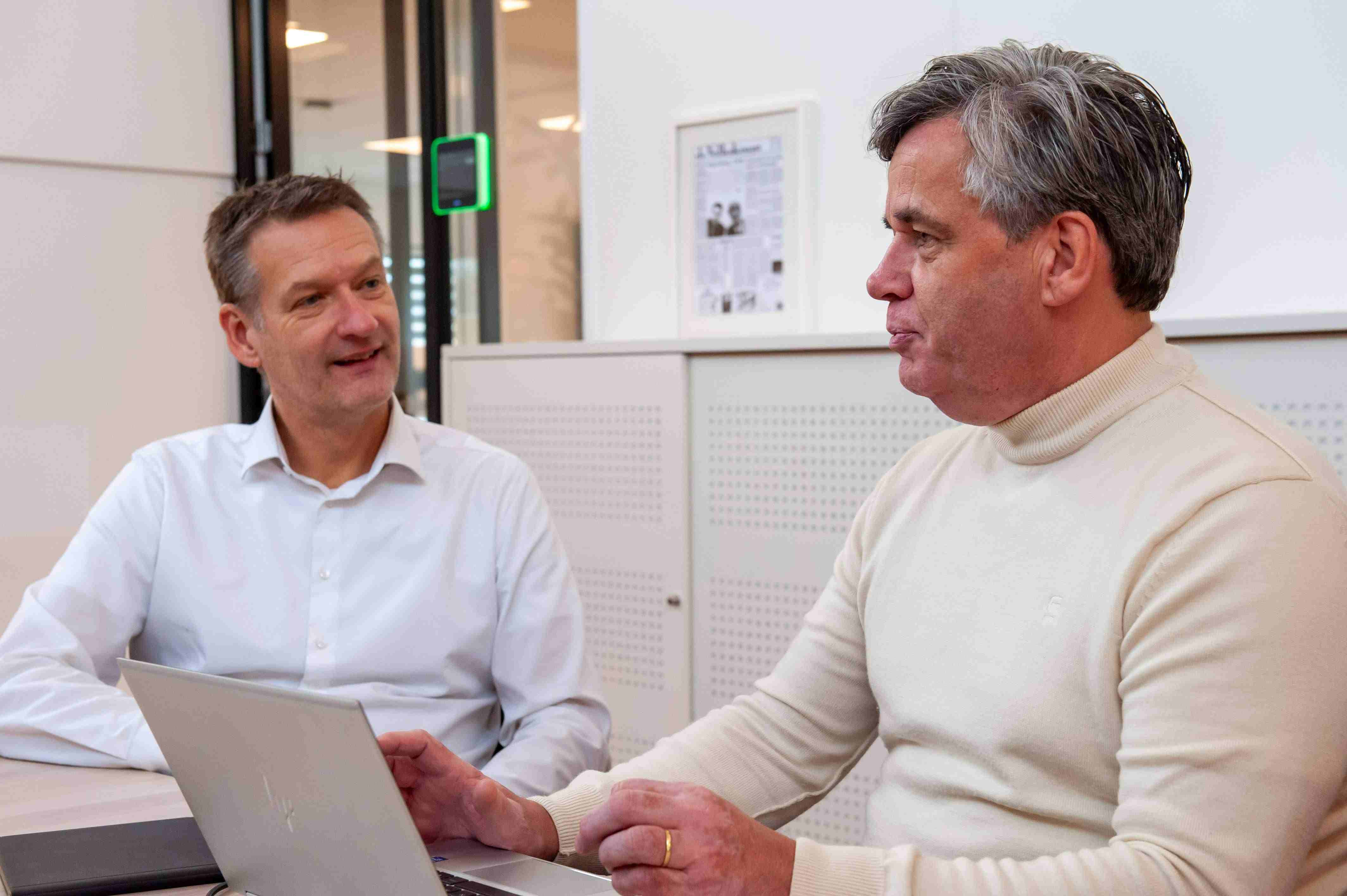 Two people talking while working on a laptop in an office.