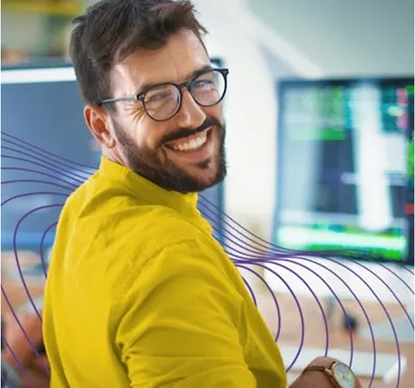 Man met een gele trui en bril die lachend over zijn schouder weg van zijn beeldscherm kijkt