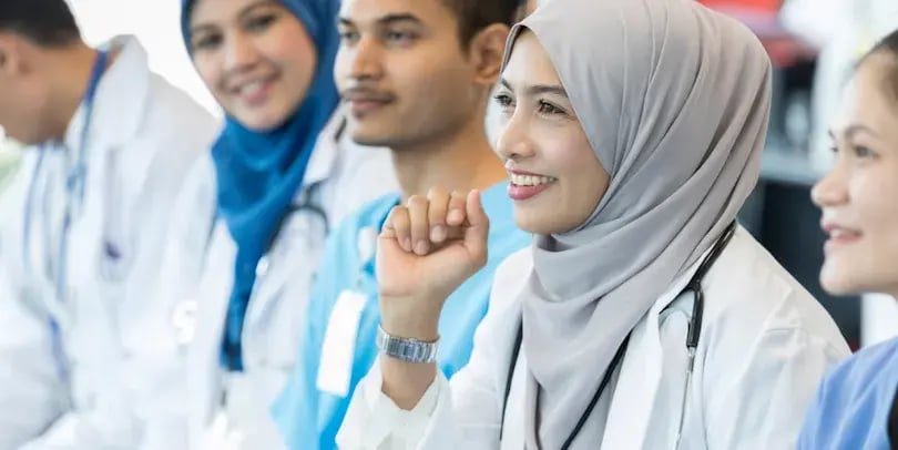 Verpleegers zitten naast elkaar, eentje met een hoofdoek heeft een vraag