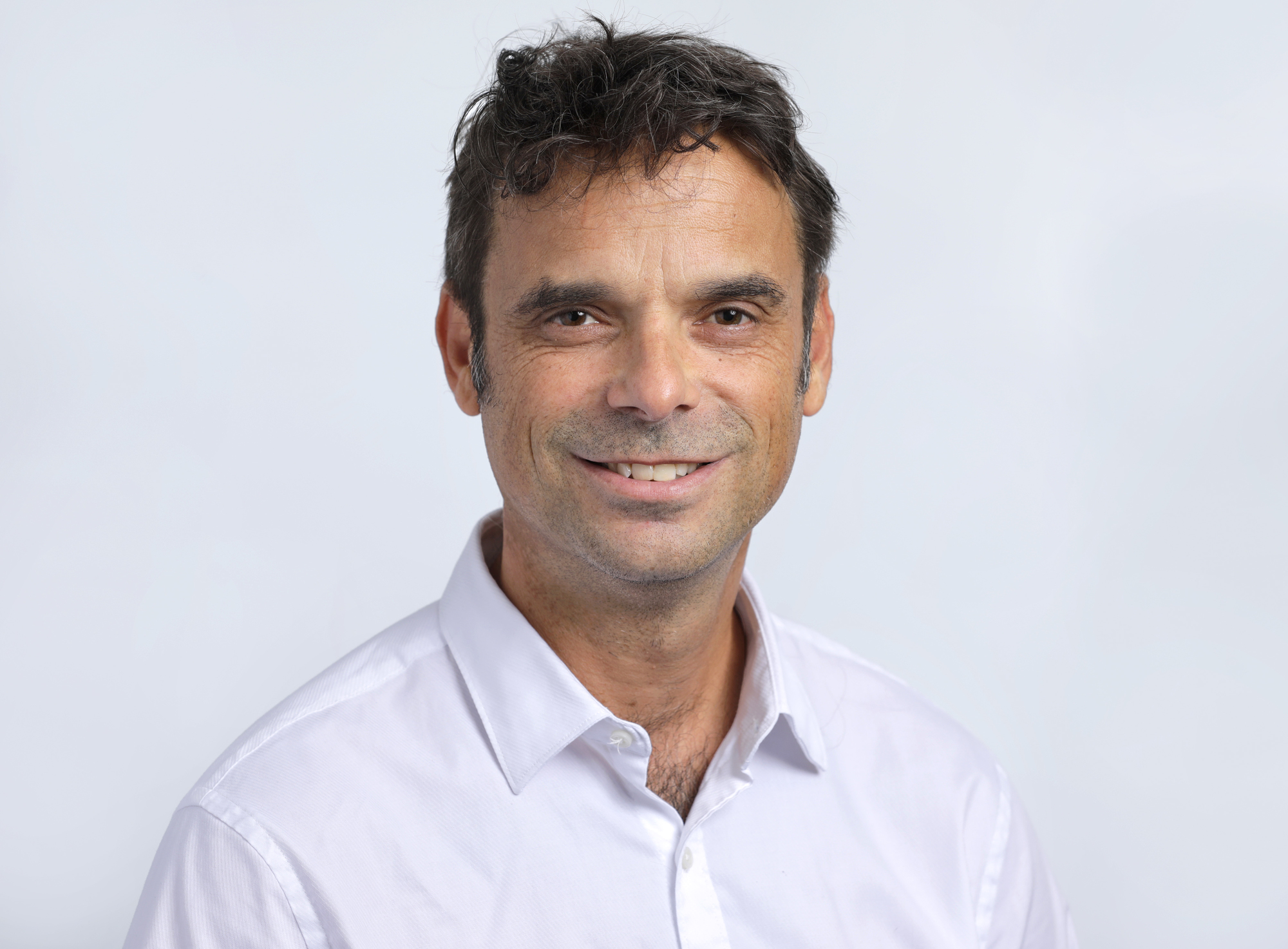 Dit is een portretfoto van een man met donkerbruin, kort haar, in een witte blouse. Hij glimlacht vriendelijk naar de camera.