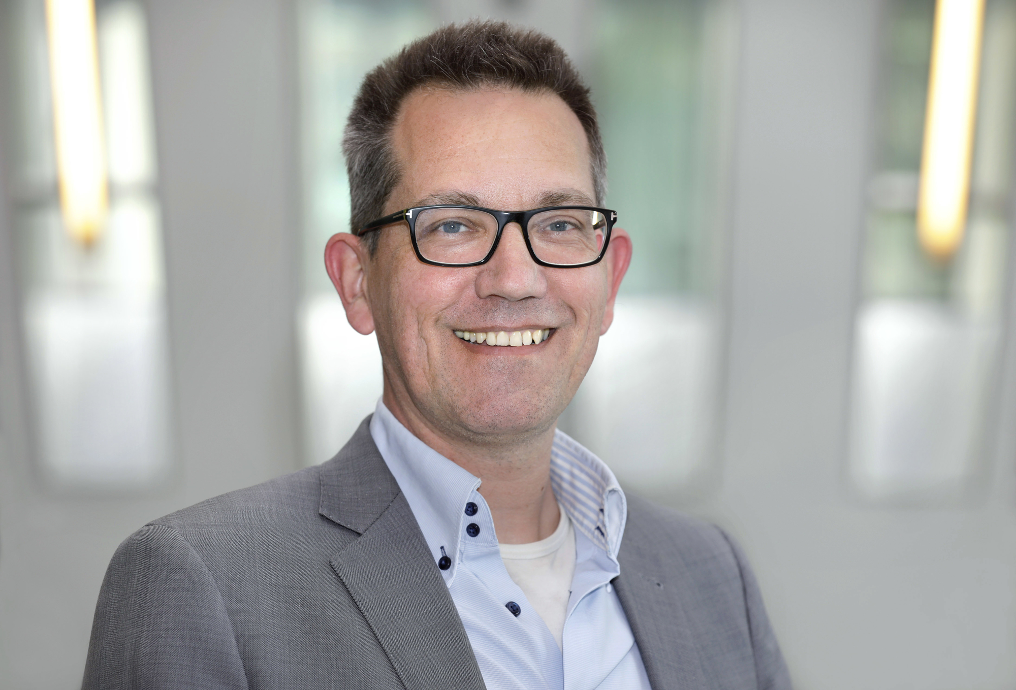 Man met bril en grijs colbert die glimlacht naar de camera, met een zachte, onscherpe achtergrond in lichtgrijze tinten.