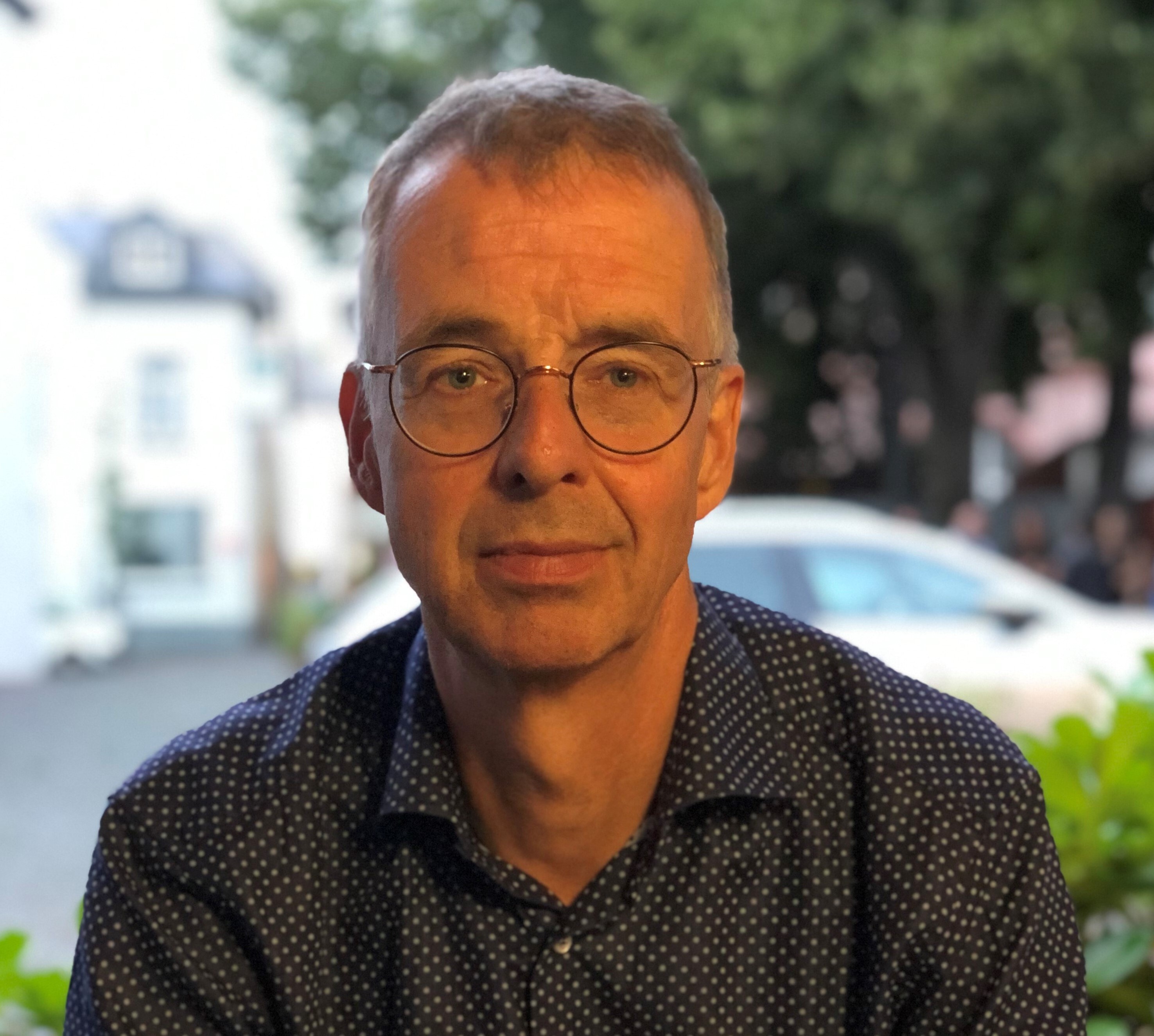 Man met bril kijkt in de camera, zittend buiten met een vage straat en auto op de achtergrond.