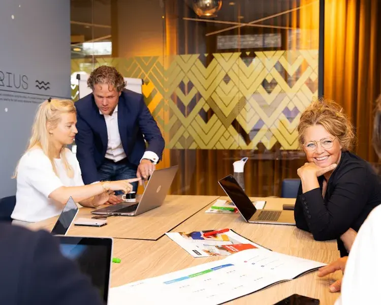 Collega's zitten aan een bureau met hun laptops en lachen en overleggen met elkaar.