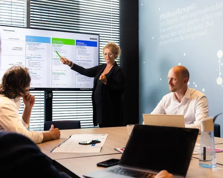 Drie mensen in een vergaderruimte en een collega wijst naar het scherm.