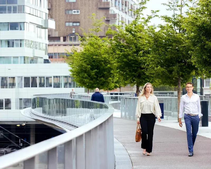 Twee mensen lopen over een moderne voetgangersbrug in een stedelijke omgeving, met bomen langs het pad en hoge gebouwen op de achtergrond.