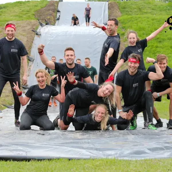 Groep sportief geklede deelnemers poseert lachend op een modderige of natte obstakelbaan, met een glijbaan op de achtergrond tijdens een actieve teamevenement.