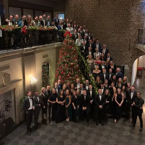 Grote groep mensen in feestelijke, formele kleding poseert samen in een historische hal rond een grote, versierde kerstboom, verdeeld over de vloer en een balkon.