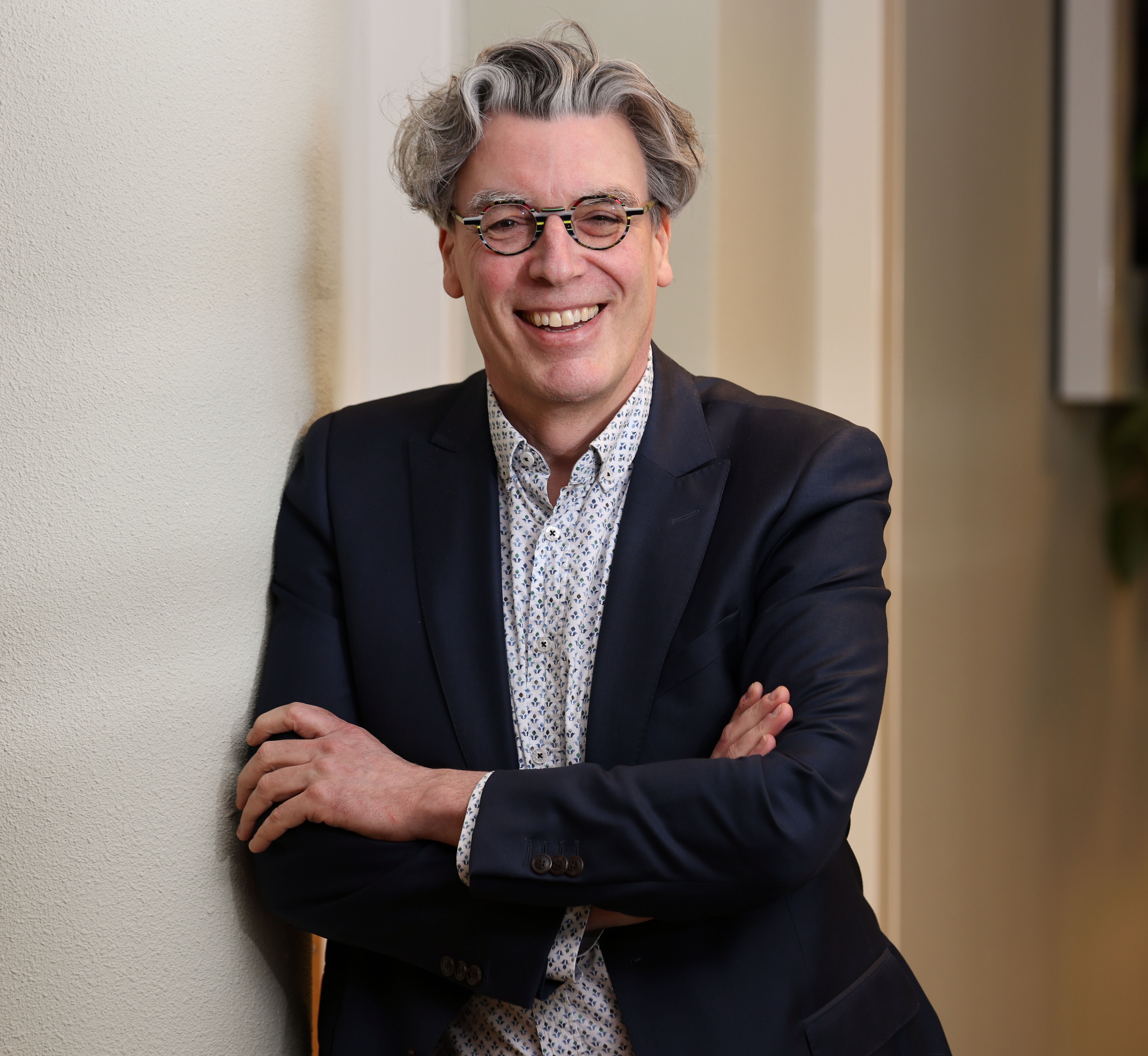 Man met bril glimlacht terwijl hij met gekruiste armen tegen een muur leunt.