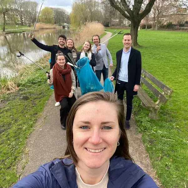 Groep mensen maakt een selfie tijdens een opruimactie in een park langs het water; ze houden afvalgrijpers en een grote afvalzak vast en glimlachen in de camera.