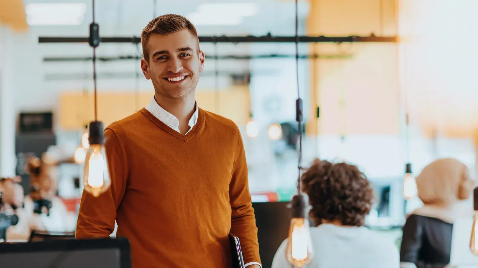 A man in an orange jumper smiles in front of him with colleagues in the background.
